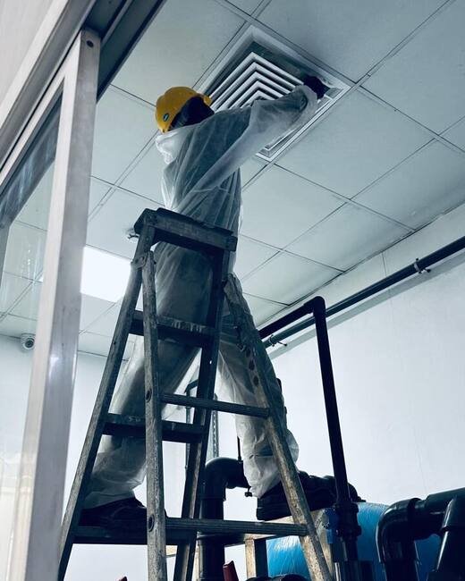 HoodEx technician in protective suit and hard hat cleaning a ceiling ventilation diffuser